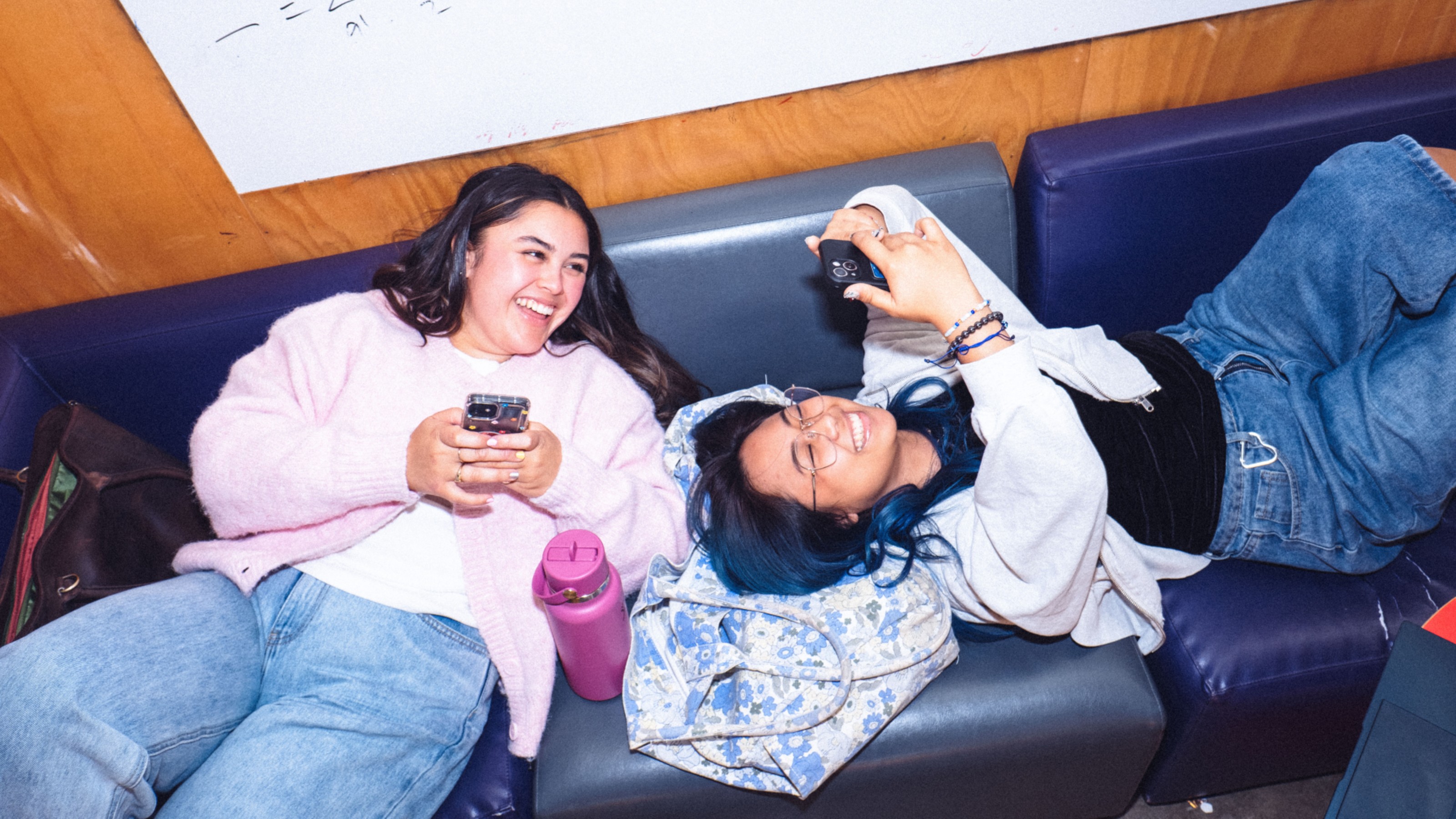 Two students relaxing on a couch sharing something on their phones.