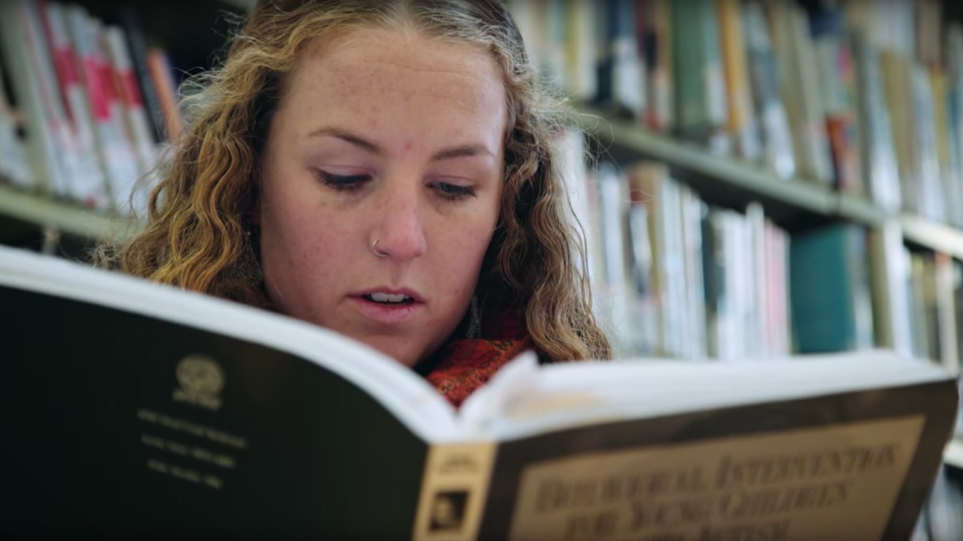A female student reads a book in front of a bookshelf.