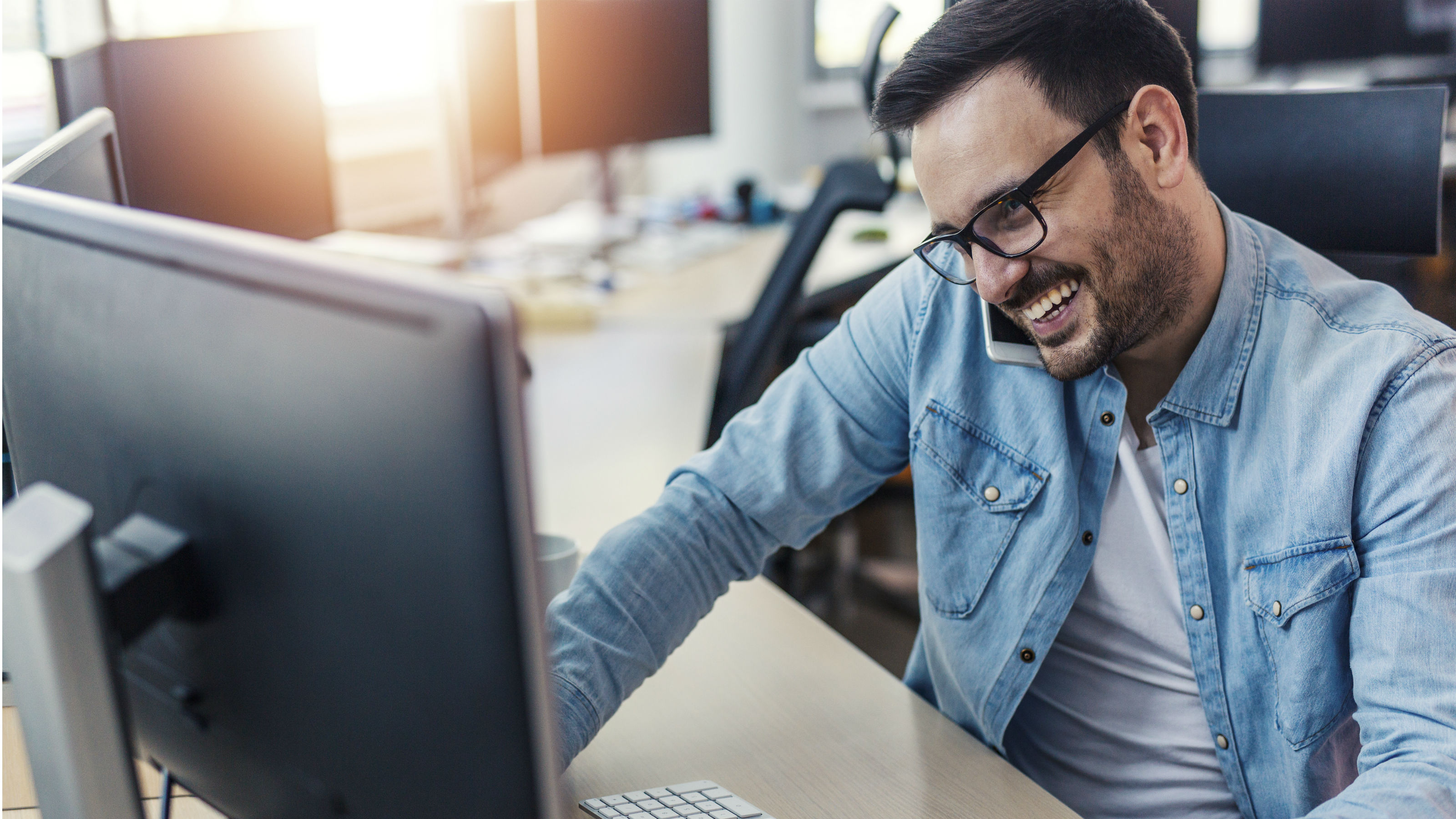 A smiling man on a smartphone, at a desk with two monitors.