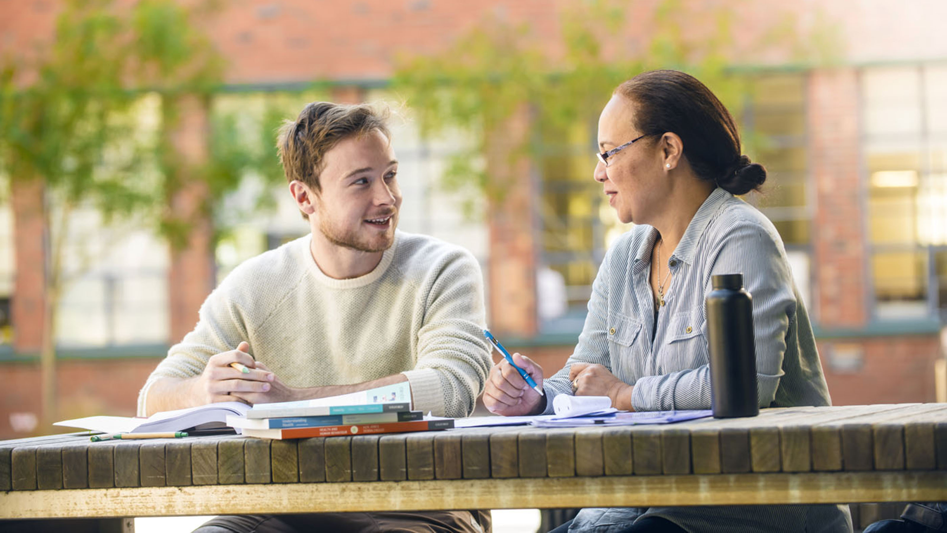 A woman and a male student with books in front of him talk while sitting outside at the University's Kelburn campus.