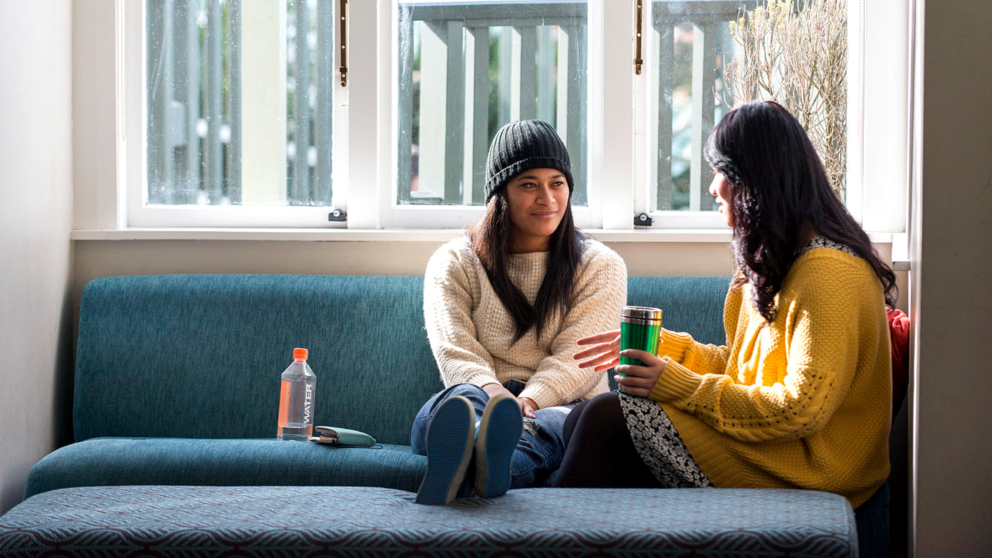 Two female students sit on a blue sofa enjoying a onversation.