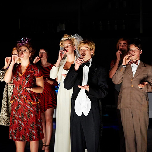 Formally dressed actresses and actors gesturing as if each of them were drinking tea from a cup in one hand and holding a saucer flat in the other.
