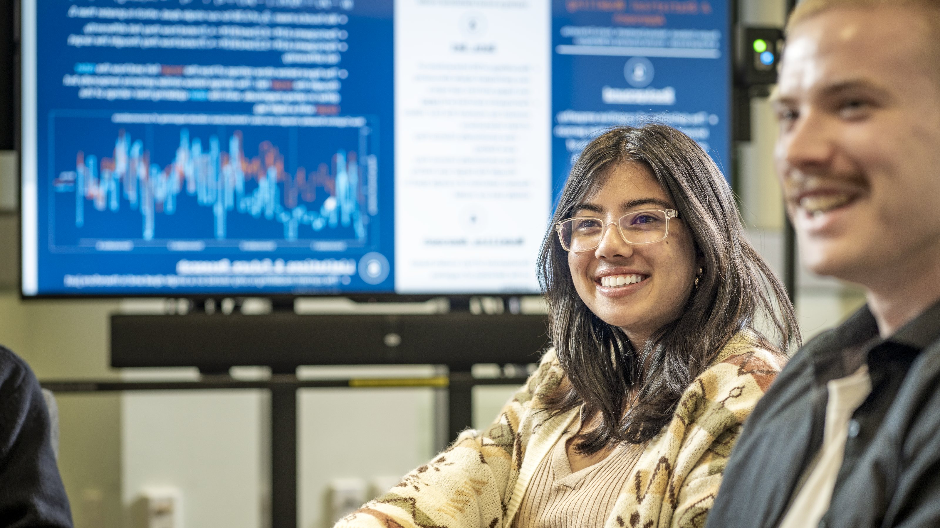 Two students smiling in a classroom setting, with a large screen behind them with (out of focus) teaching notes.