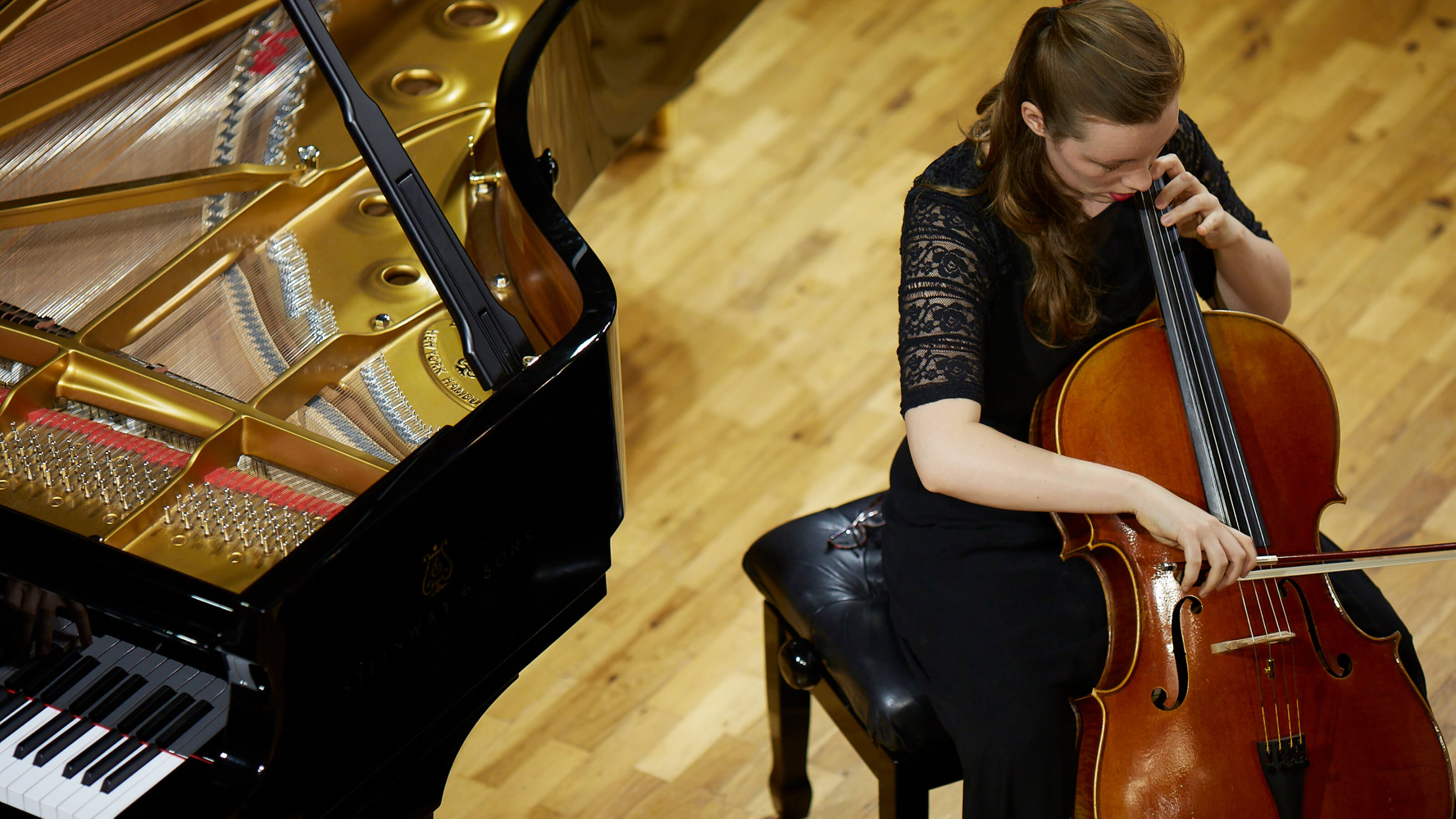 A female musician plays the cello while sitting beside a grand piano.