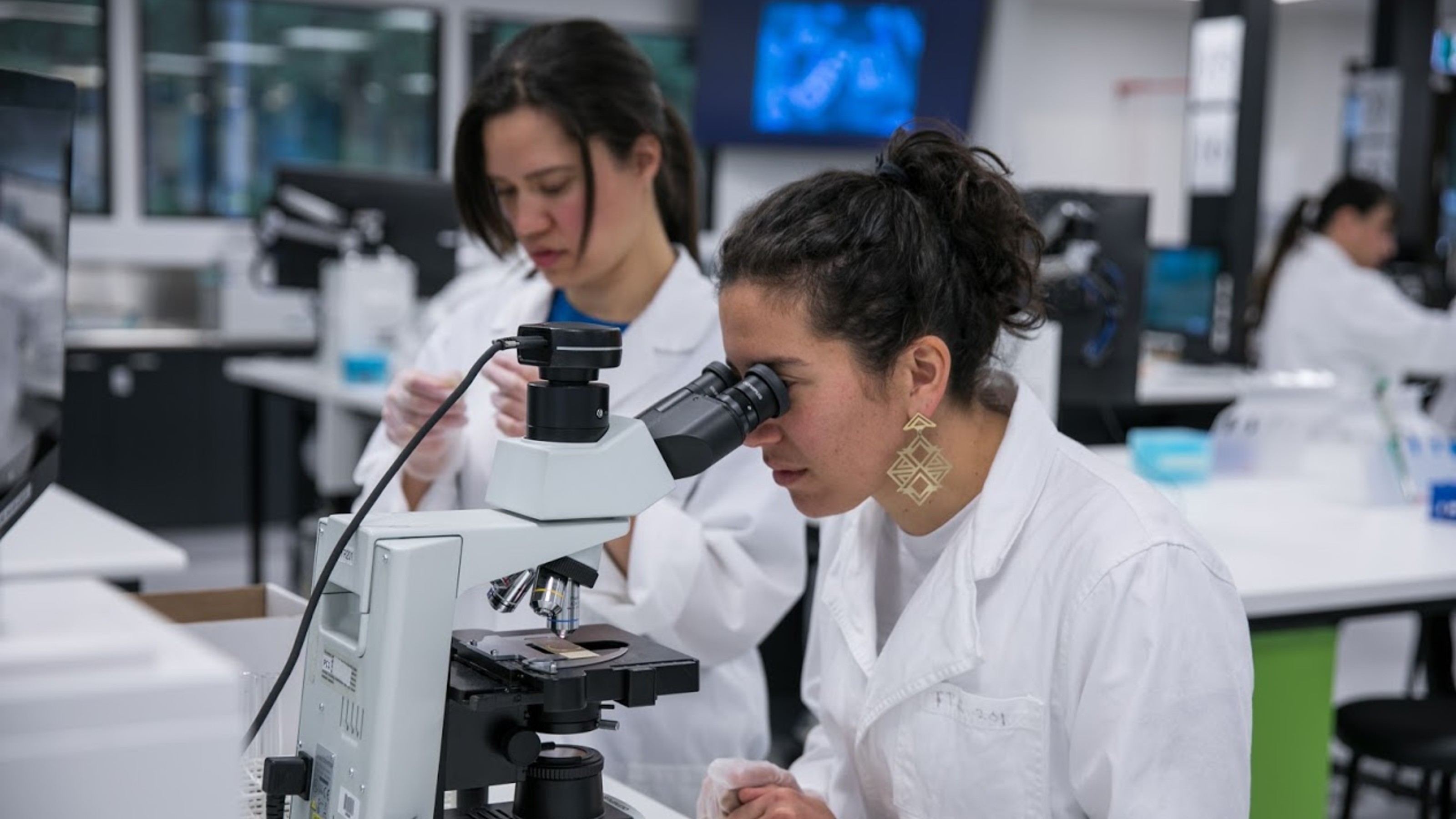 Student in white lab coat looking into a microscope.