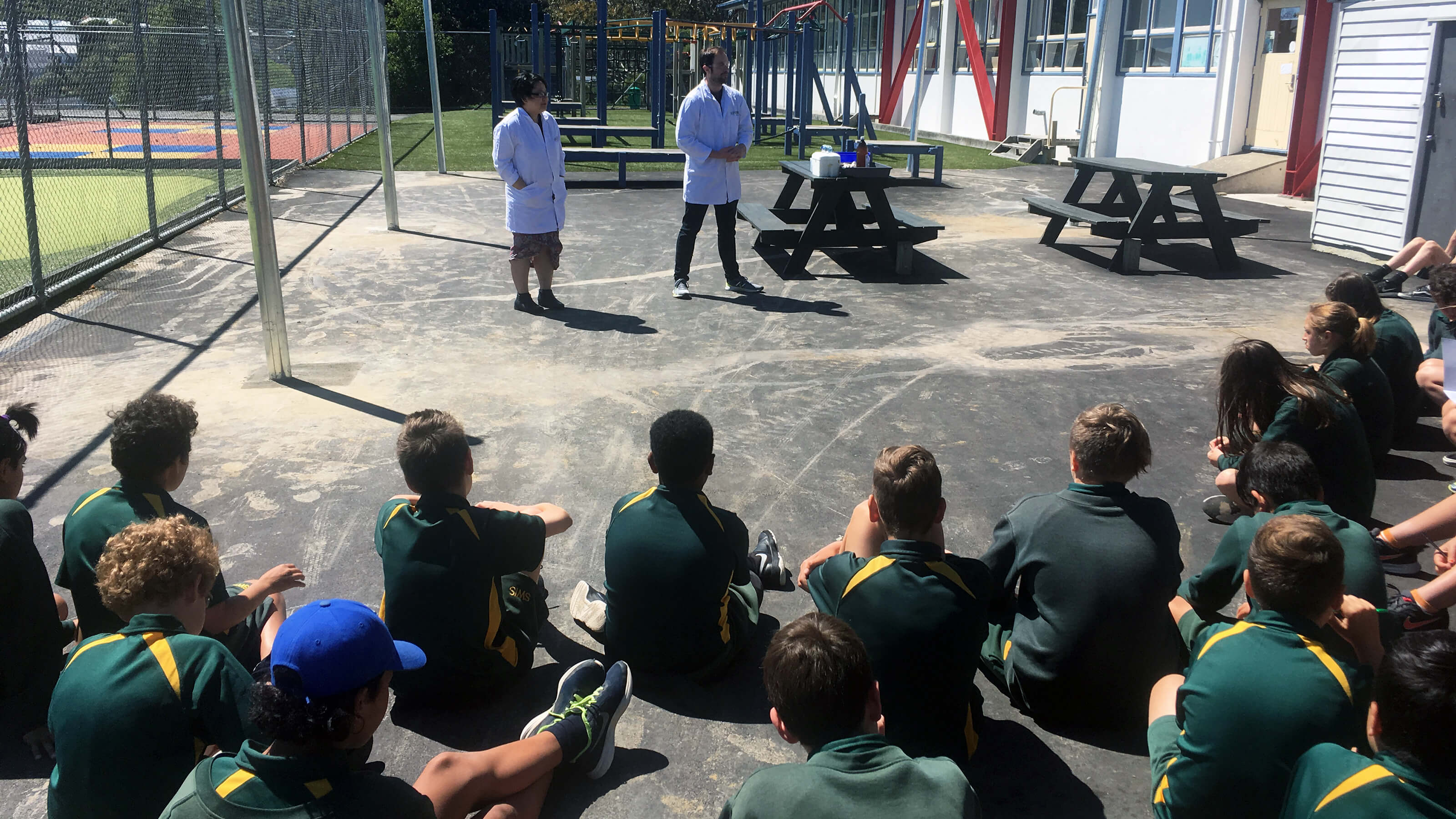 A group of students at a primary school watch two people in lab coats perform a science experiment.