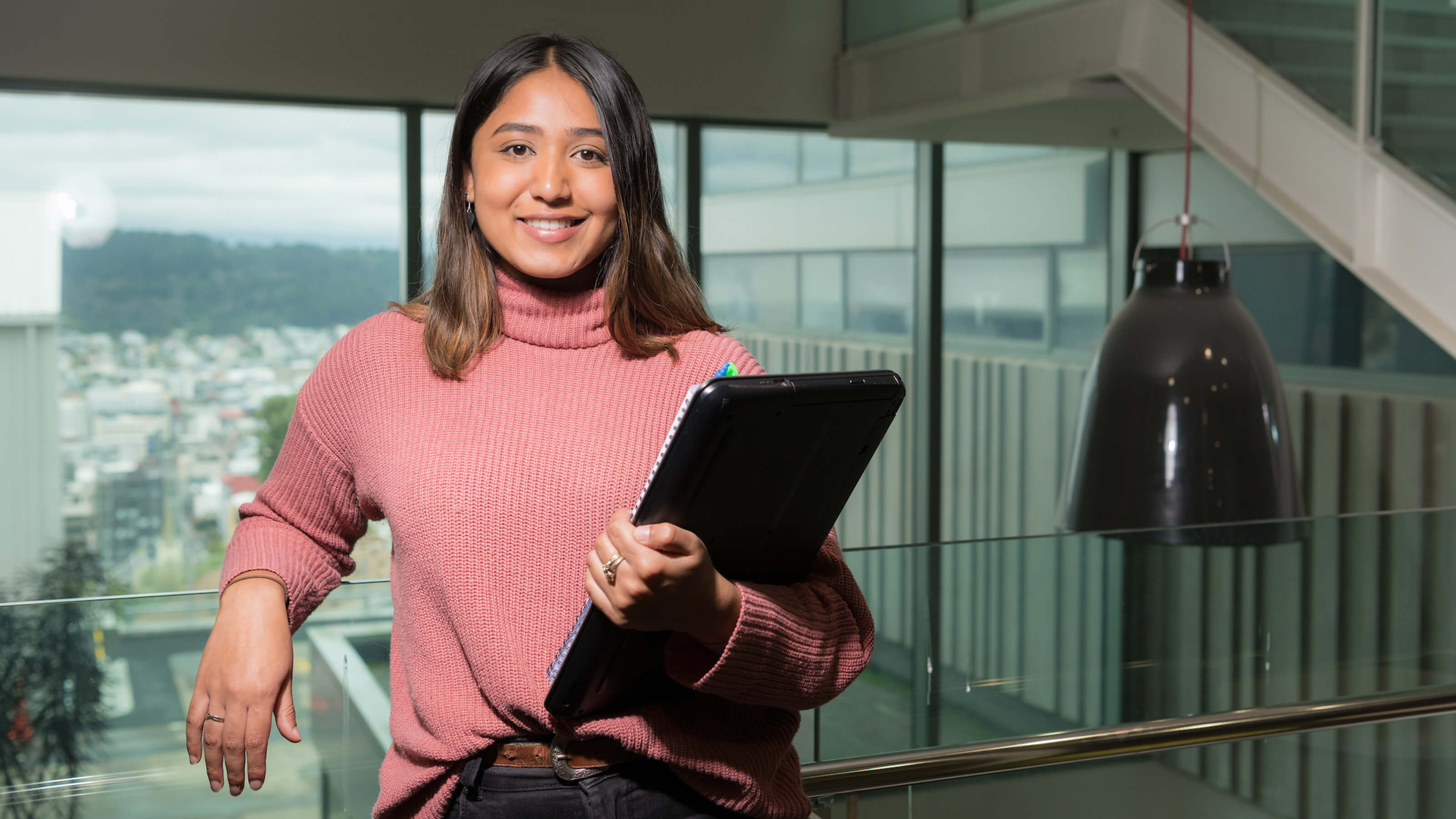 Law student Komal standing with laptop and books in front of Wellington view.