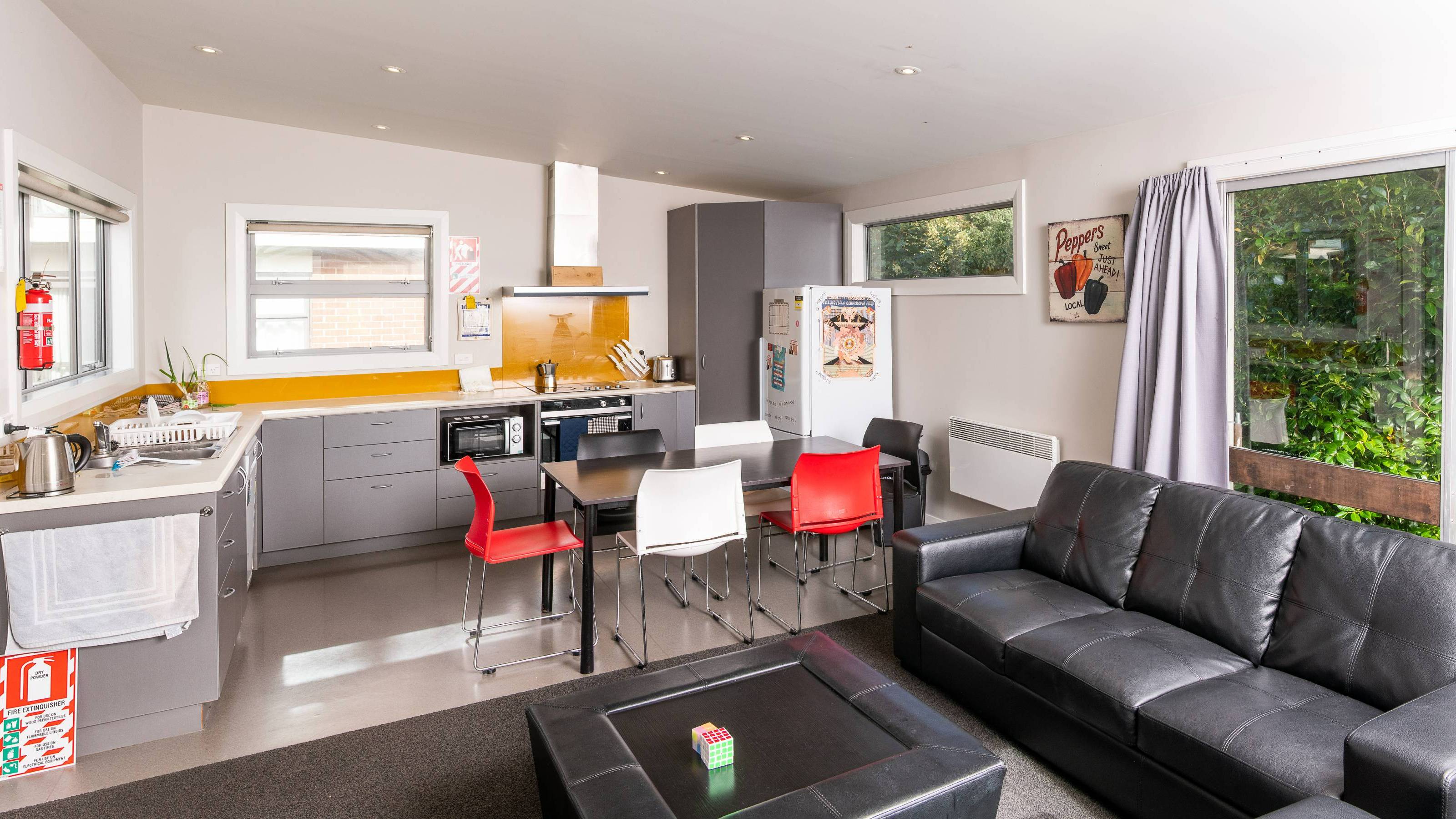 An open plan kitchen/living room with black leather couches and coffee table, grey kitchen counters, and a black dining table with red, white, and black chairs.