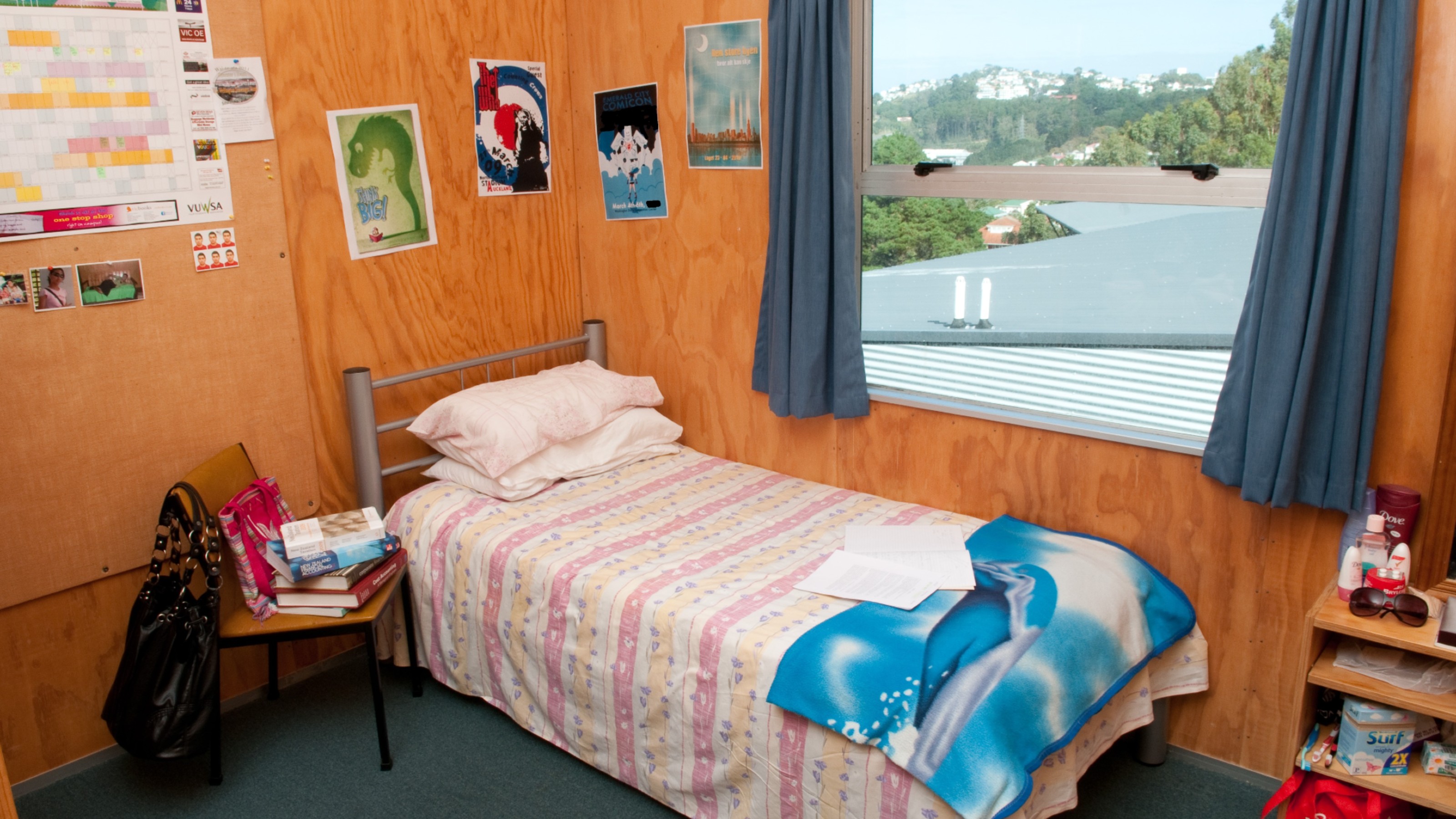 Bedroom shows single bed, chair, shelves, posters and calendar on walls, and view of forested hills across the city.