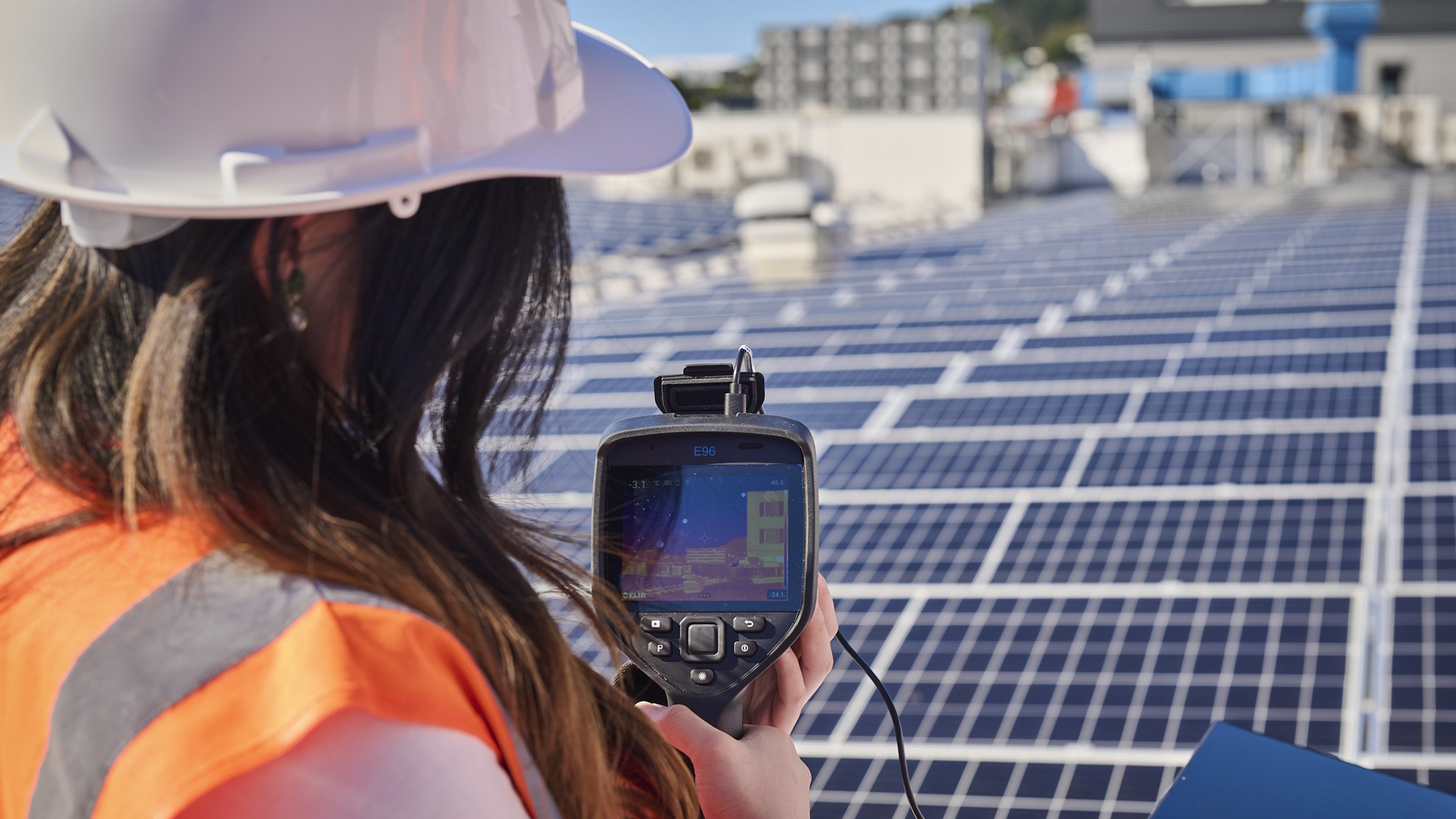 Person in hard hat holding measuring device in front of rooftop array of solar panels.