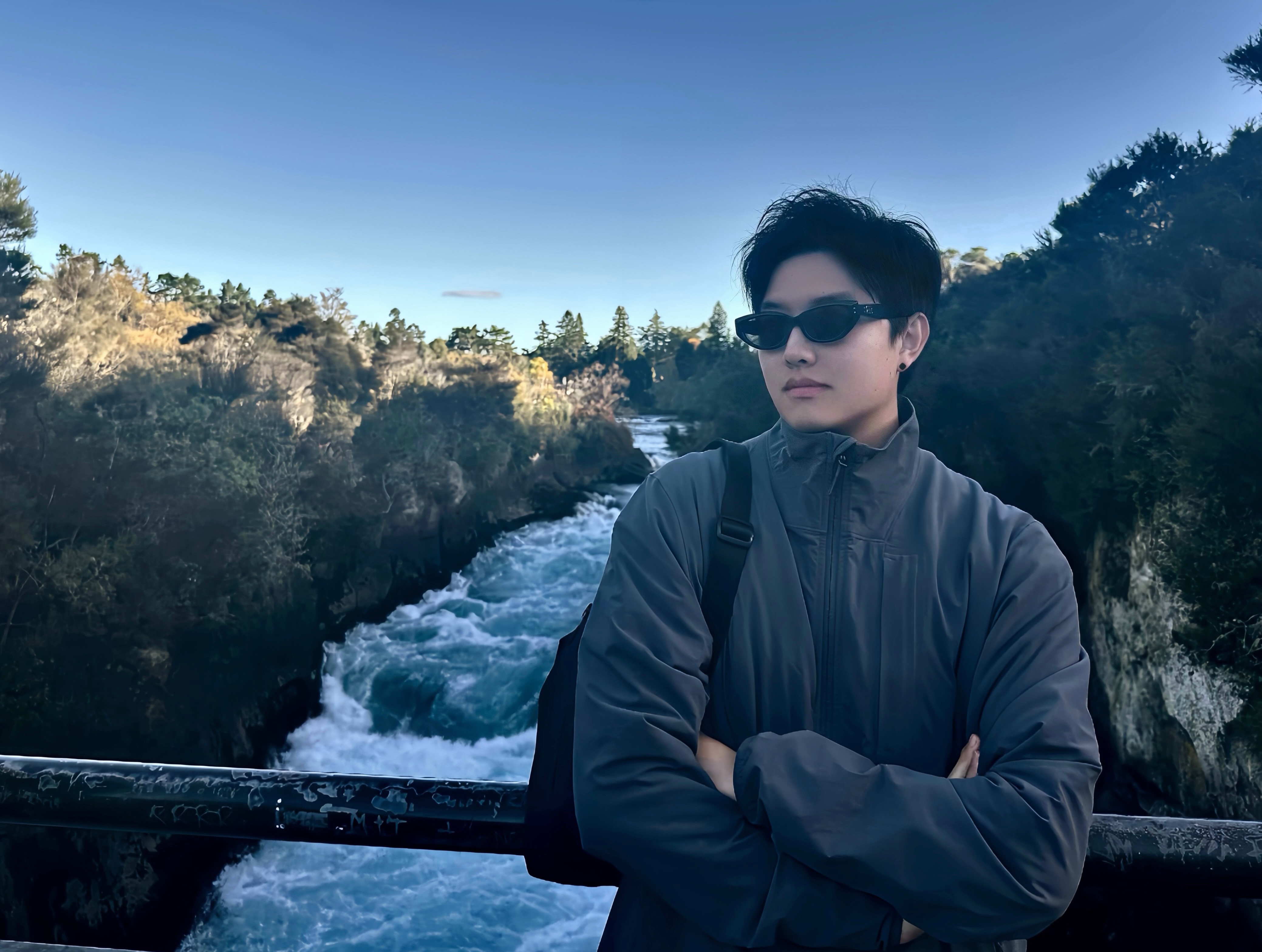 A person in a gray jacket and black sunglasses stands on a bridge, arms crossed, looking to the right. They wear a black backpack over one shoulder. Below, a turbulent river with white water flows between forested banks, under a blue sky with scattered clouds.