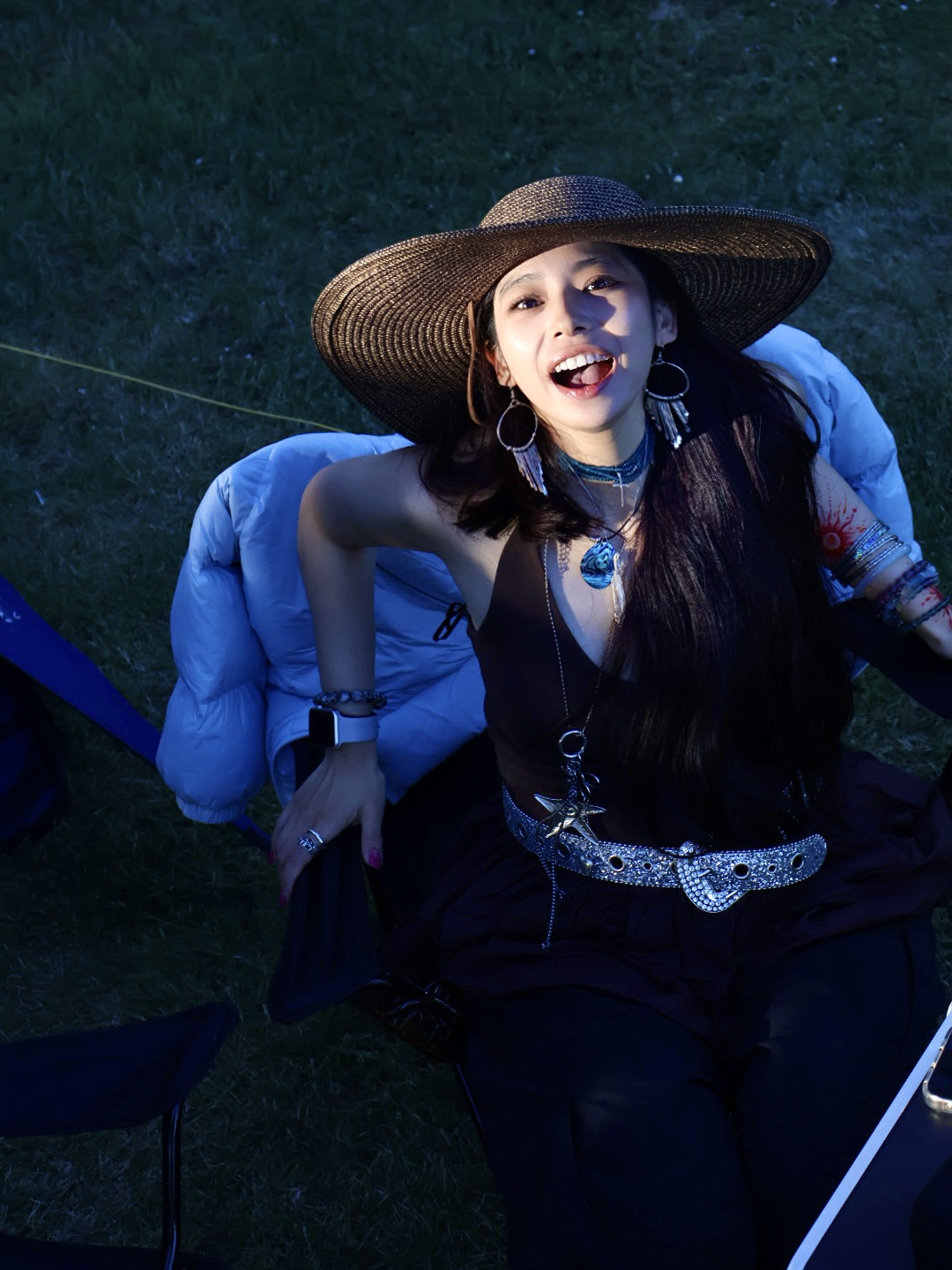 woman with tan skin and dark hair, wearing a wide-brimmed hat, visible jewelry (such as earrings or a necklace), and a dark-colored outfit. She is seated on a grassy outdoor surface, with a folded or draped jacket placed behind her. Her expression is playful and engaging, and she is looking upward—directly at the camera—with warmth and confidence. The setting appears to be a sunlit, natural environment.