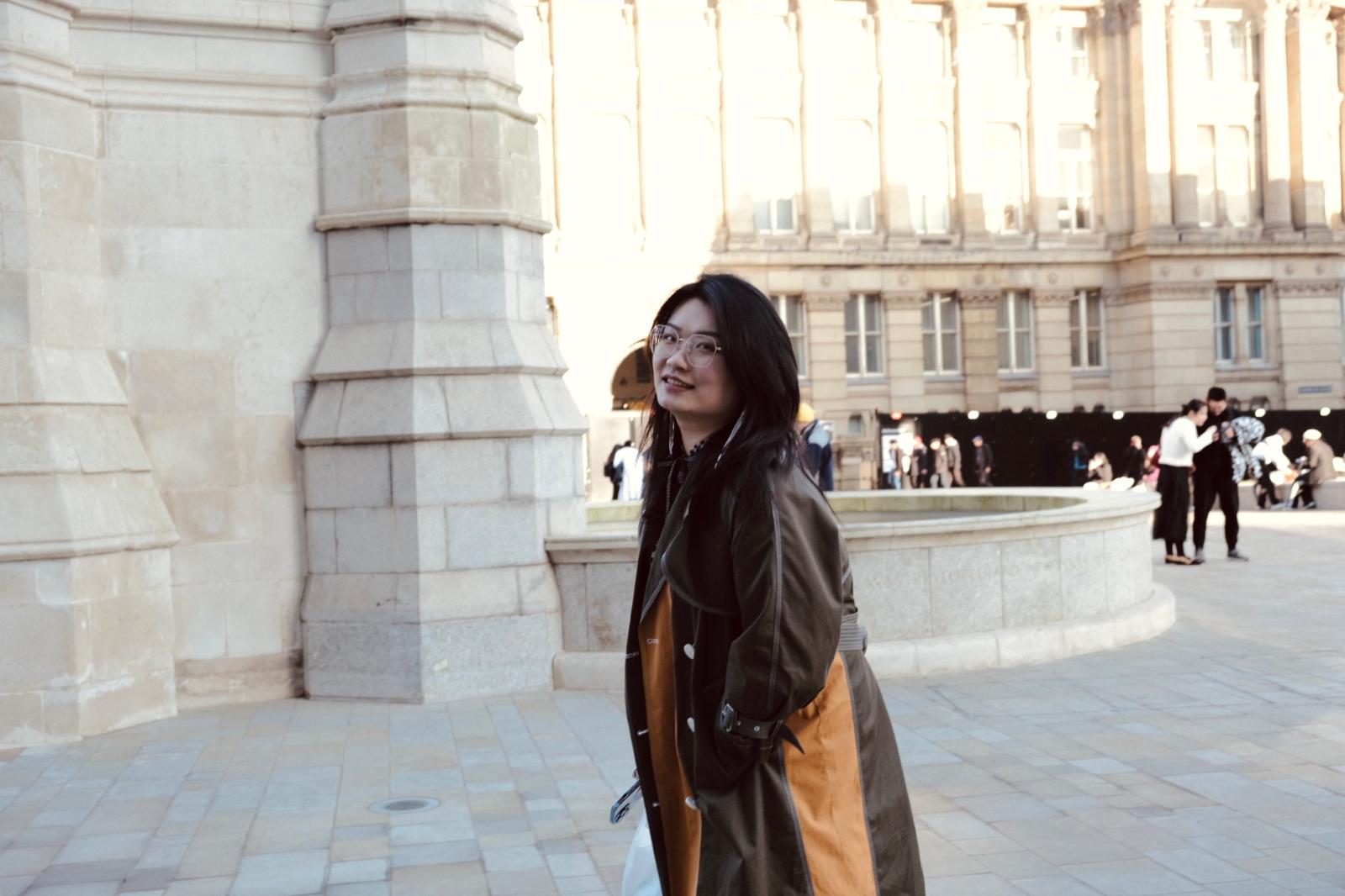 A woman with short black hair and glasses stands in a public square wearing a long two-tone trench coat—dark green on one side, orange on the other. Behind her rises a large stone building with columns and multiple windows; other people walk in the background.