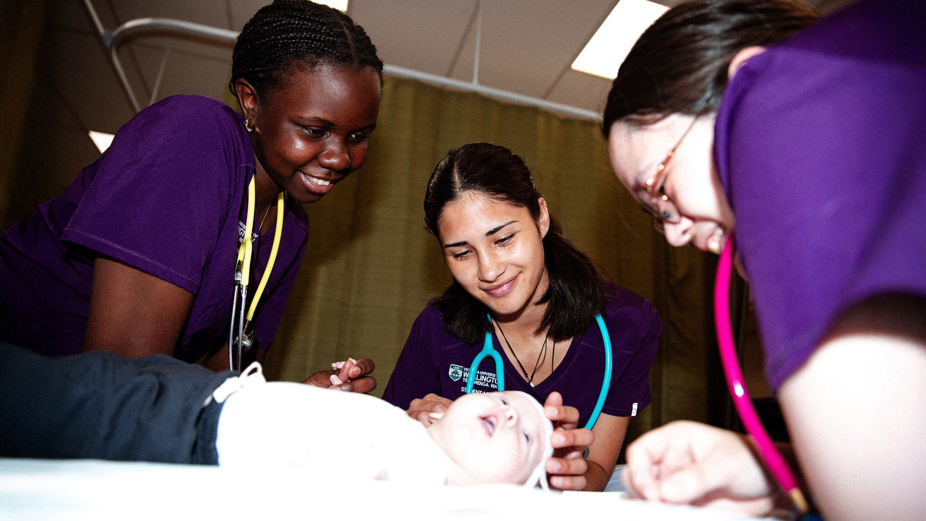 students in purple scrubs with stethoscope examining a small baby