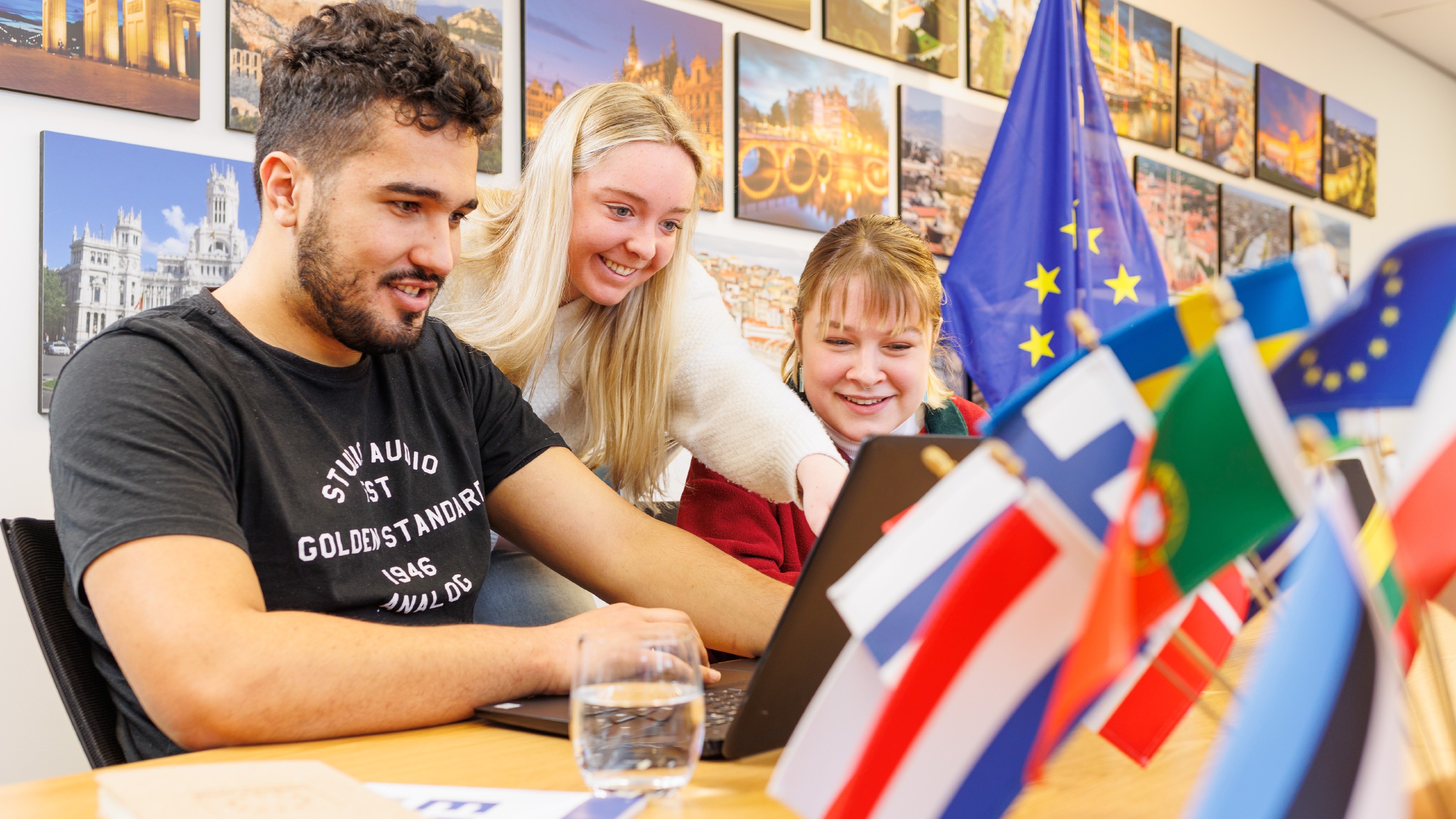 Three students sitting around a laptop, surrounded by colourful flags