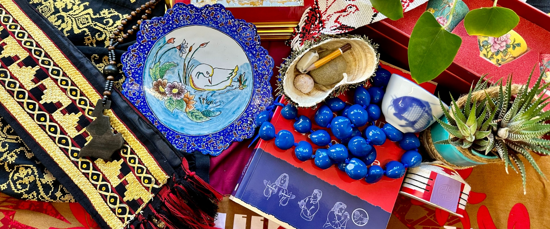A colourful display of cultural items including embroidered textiles, a painted ceramic plate, traditional jewellery, beaded crafts, shells, plants, and ornate containers.