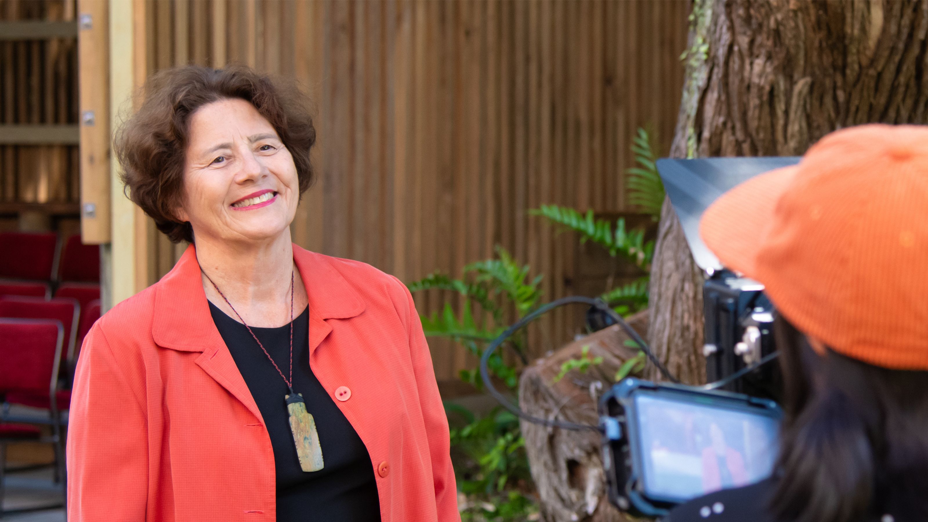 Woman wearing a red jacket smiling in a relaxed way, with a camera person with red hat in the foreground, wooden building in the background