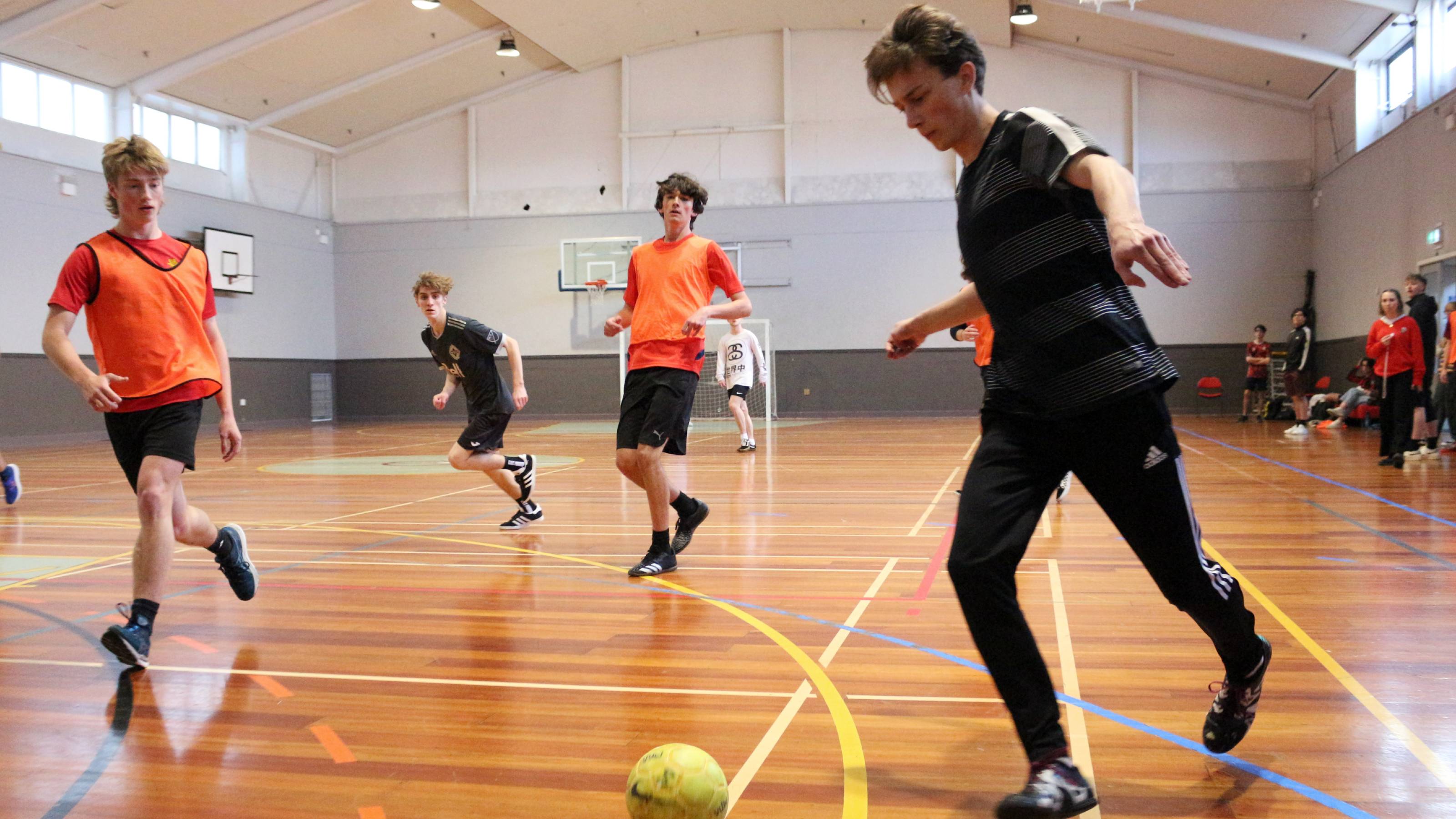 A player dressed in black dribbles in futsal, hotly pursued by players in orange