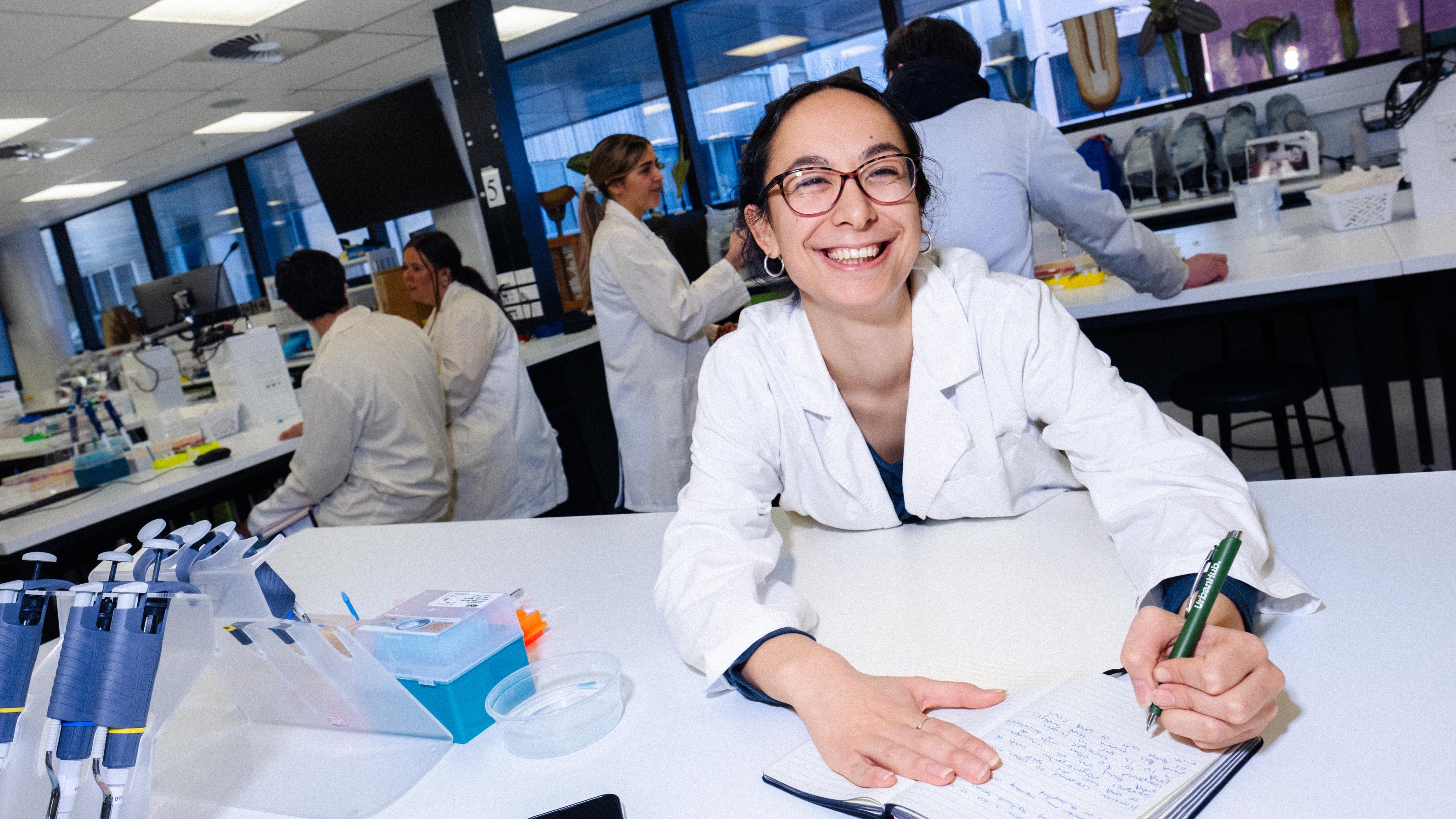 Student in a lab setting with other students in the background, wearing a white lab coat and writing in a notebook