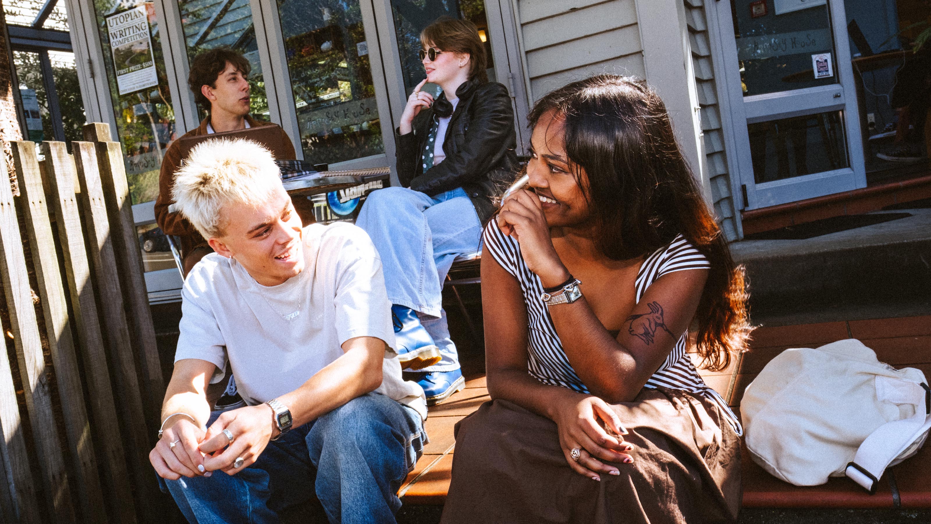 2 students sitting on stairs outside a building, with 2 other students sitting behind them at a table