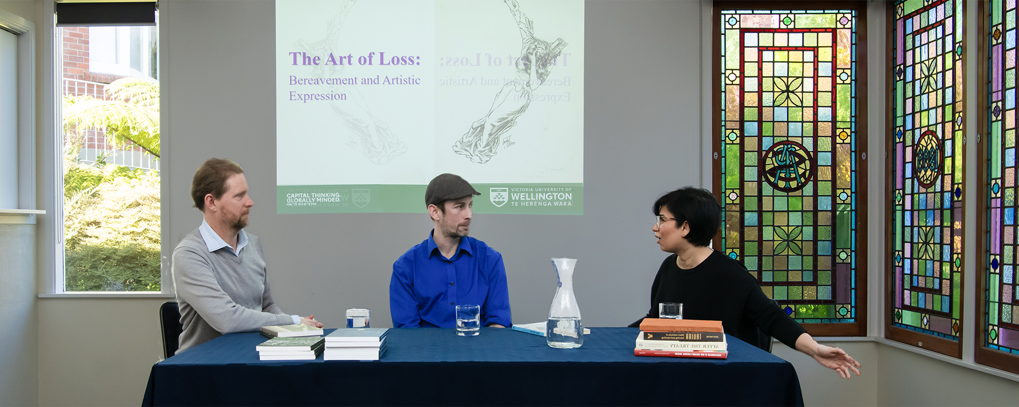 Two men and one woman sitting at a desk. They have water and books sitting in front of them. The woman is talking and gesticulating with her hands.