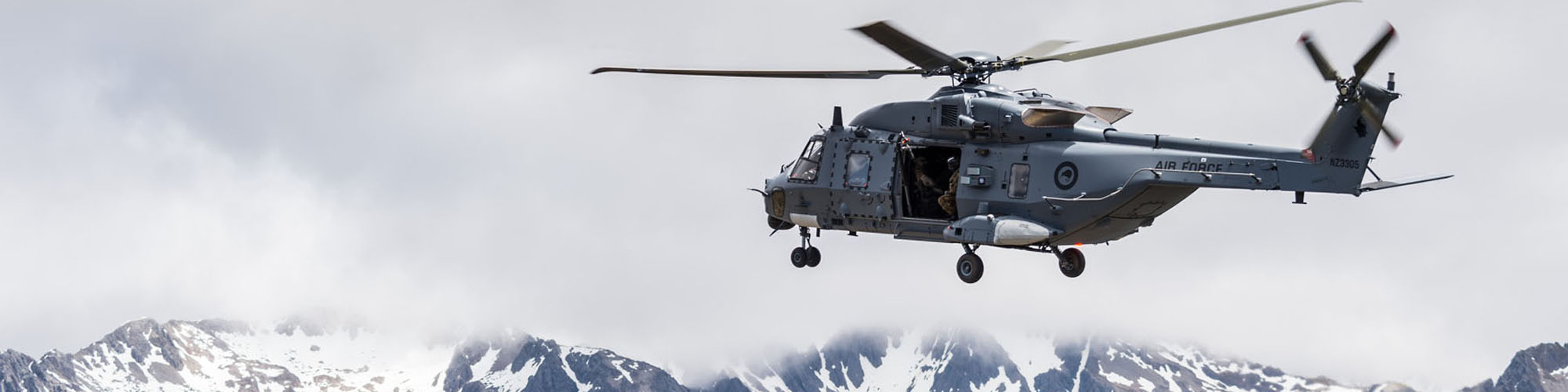 A large military helicopter hovers in front of mountains and clouds.