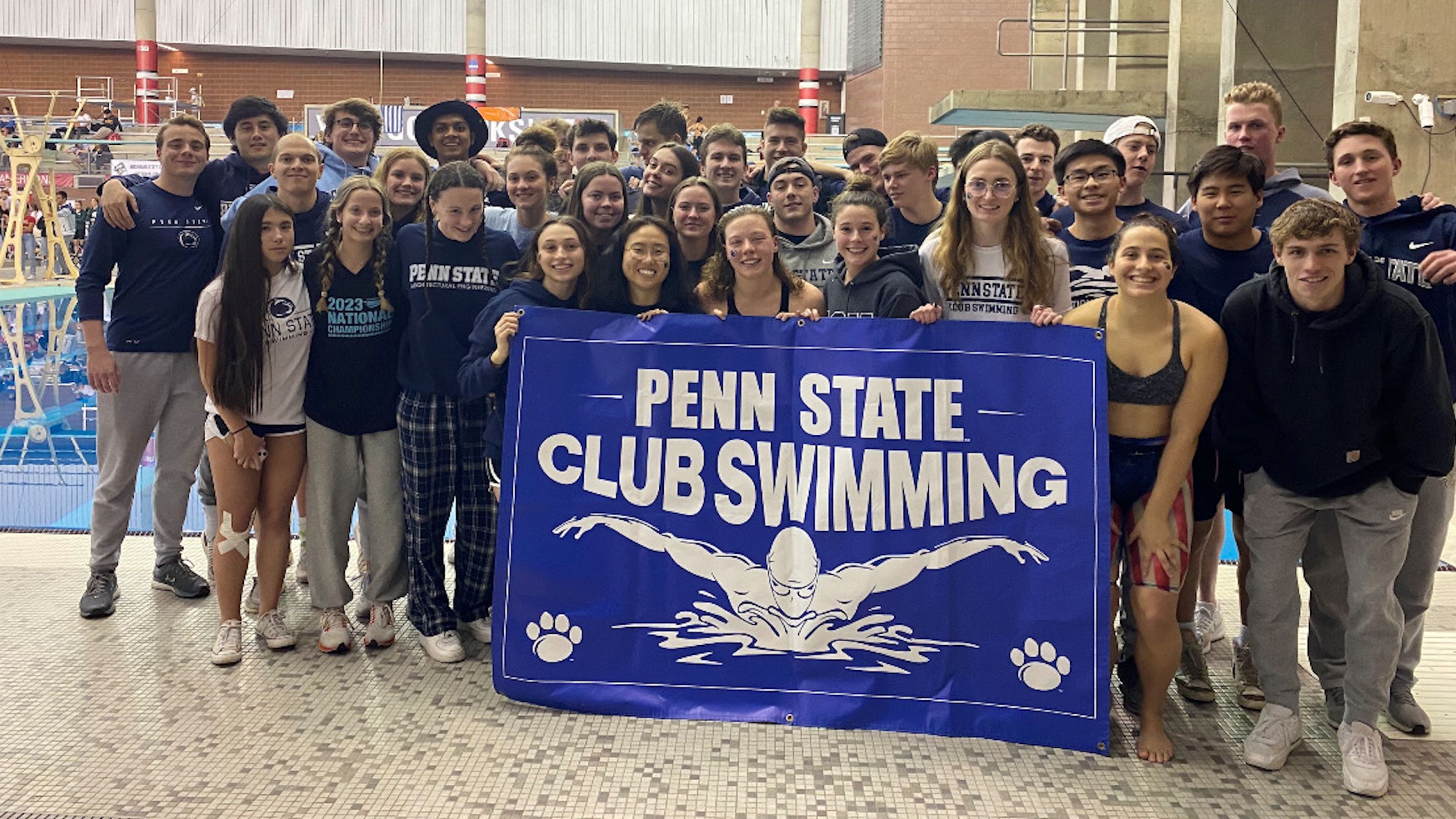 A group of students standing in front of a swimming pool, holding a larger banner that reads 'Penn state club swimming'.