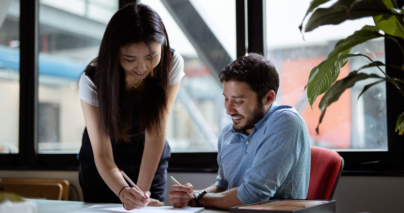 A student standing explains work to another student seated at a table, with a plant and window in the background.